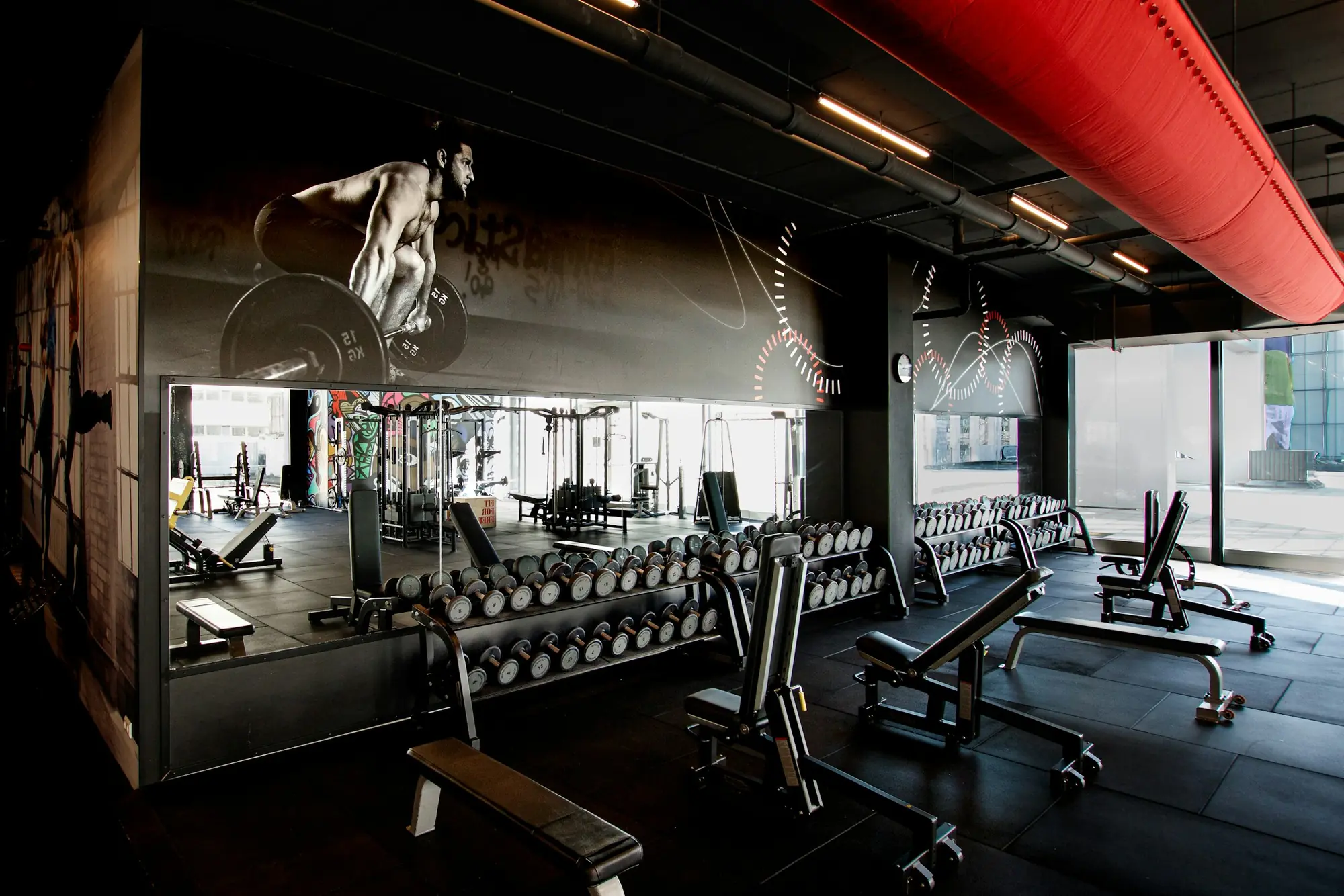 A dark gym floor with rows of dumbbells and benches under a red duct